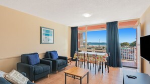 En stue med sofa, sofabord og spiseområde med bord og stole. Der er et stort vindue med udsigt til havet og bygninger.