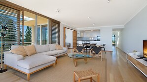 A modern living room with a sofa, coffee table, and chairs, a kitchen in the background, and large windows with blinds. - Orion Unit 7 Modern apartment with Wi-Fi included, overlooking Snapper Rocks and Rainbow Bay (Coolangatta)