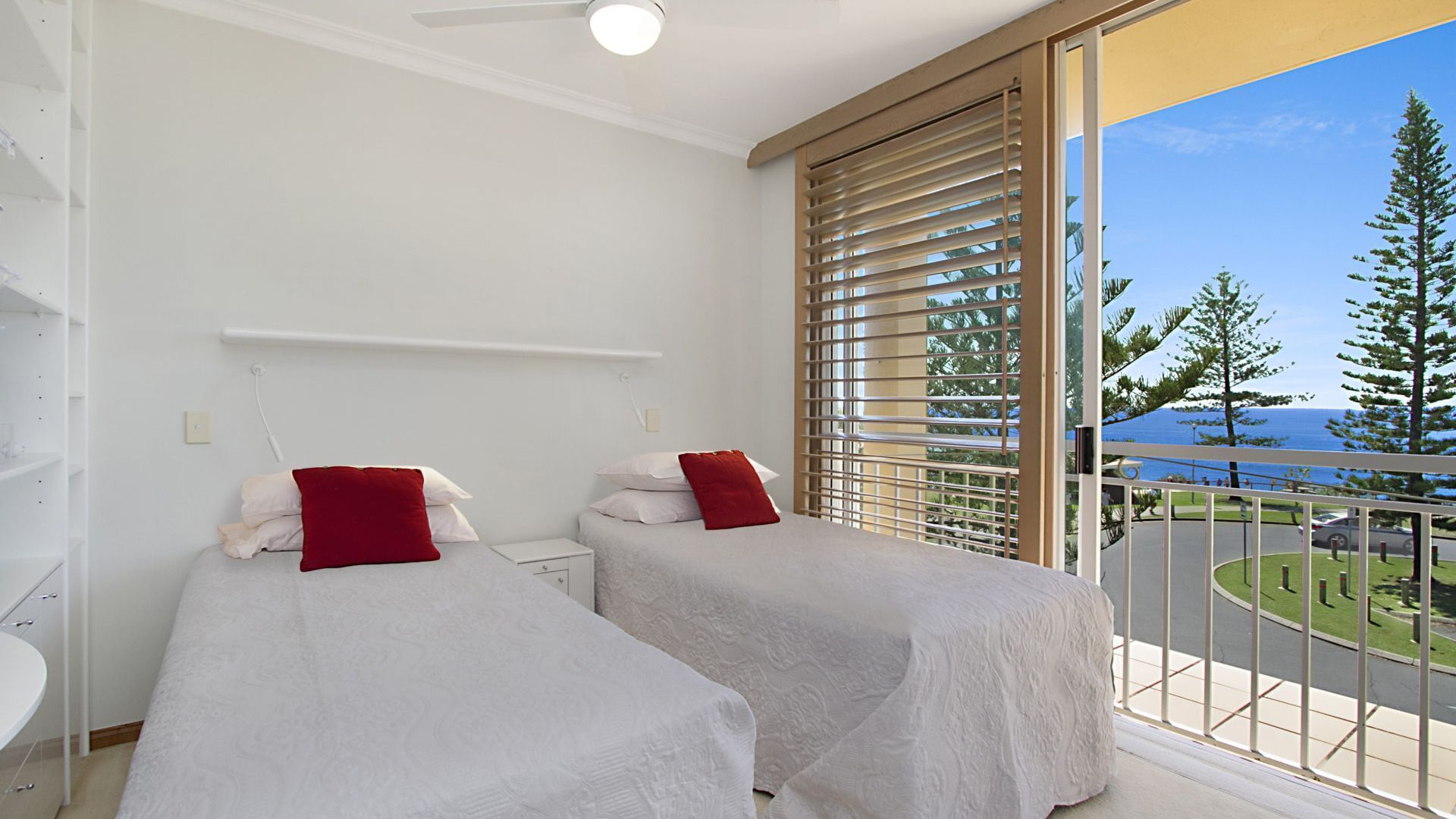 A bedroom with two beds, white bedding, and red pillows. There is a window with blinds, a white ceiling fan, and a view of a coastal area with trees and a road.