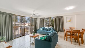 A spacious living room with a blue sofa, a dining table with chairs, and large windows with curtains. - Cobden Court Unit 6 Walking distance to Rainbow Bay and Snapper Rocks beaches with Wi-Fi Inc. (Coolangatta)