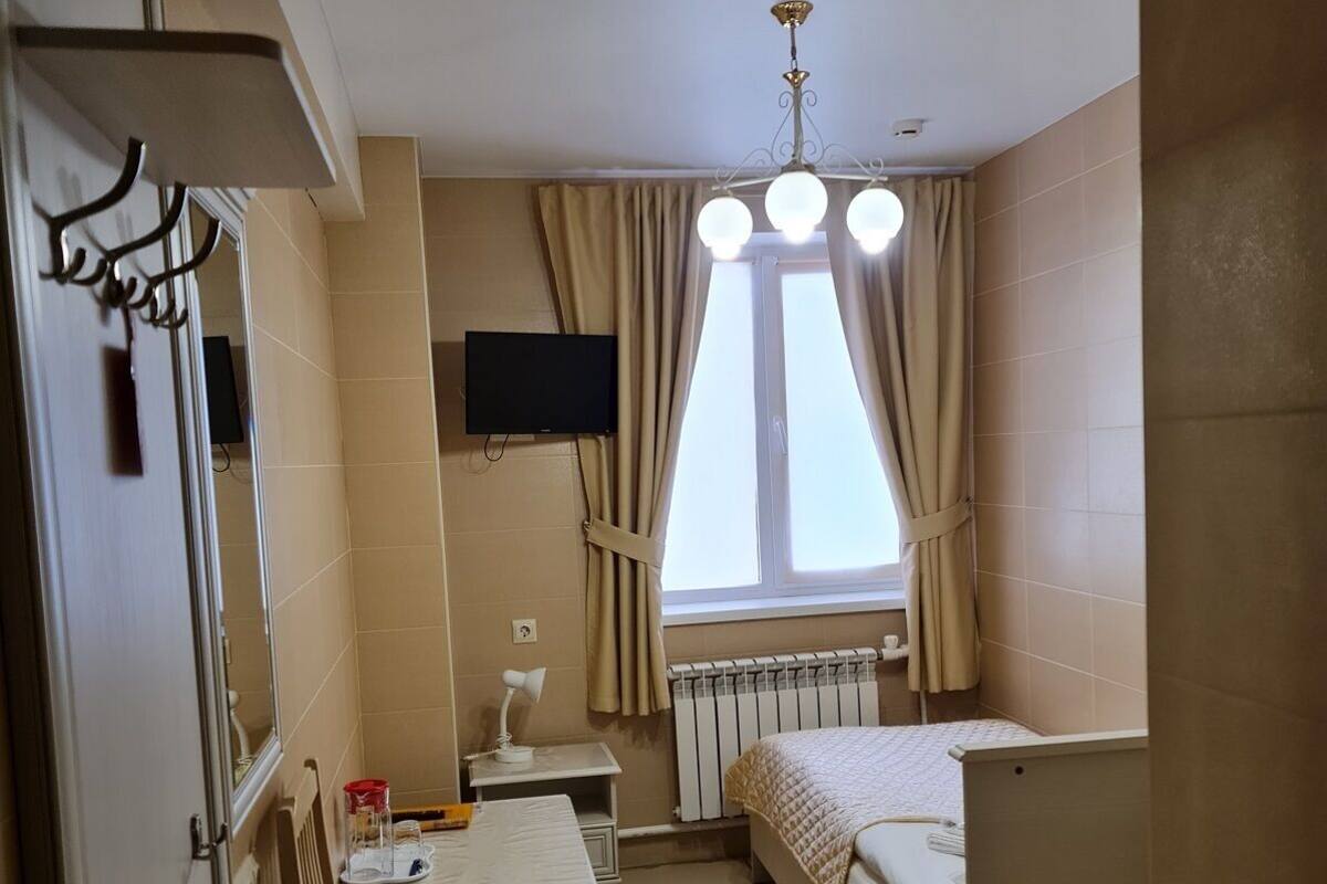Une petite pièce bien éclairée, avec un lit, un bureau et une chaise, un téléviseur fixé au mur et une fenêtre avec des rideaux.