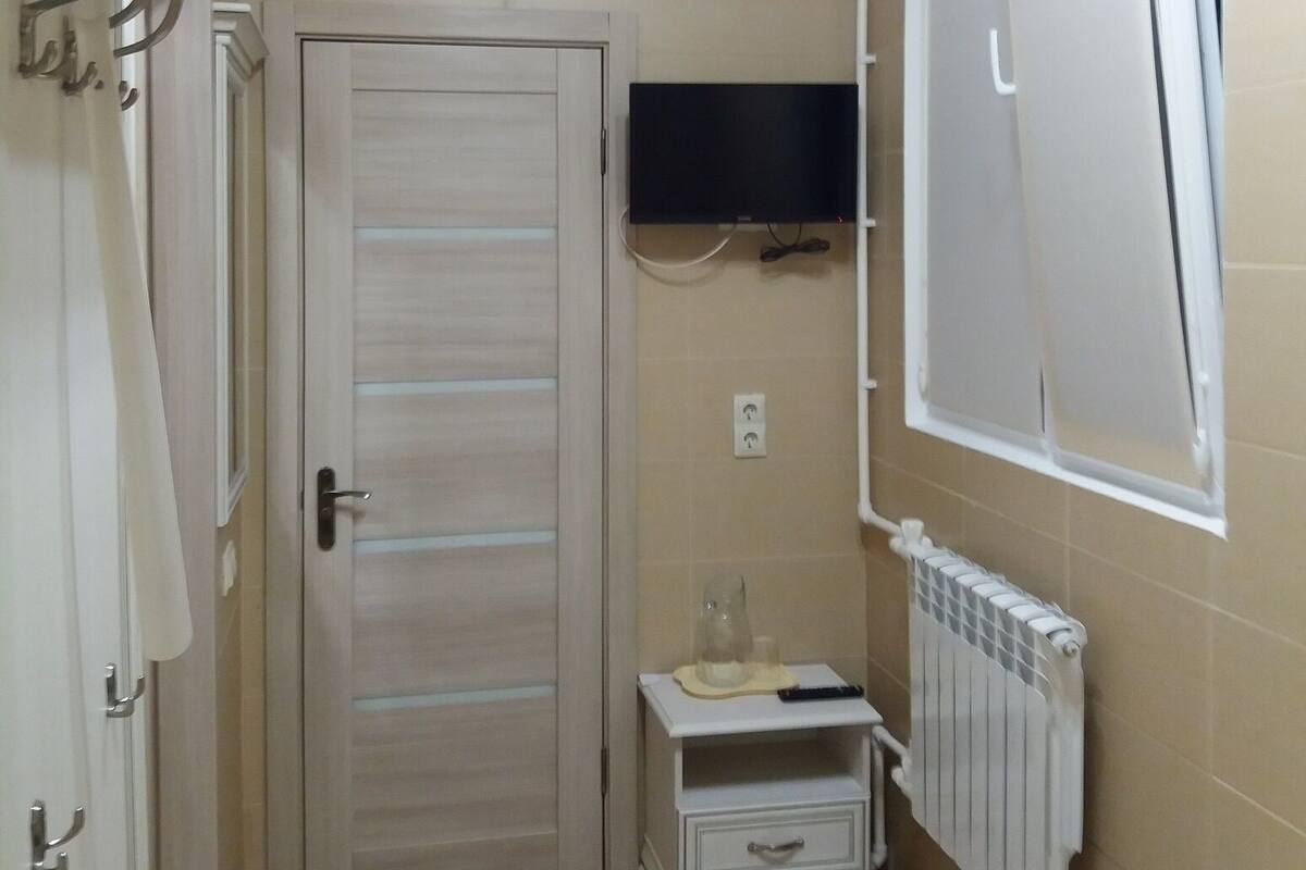 Une petite pièce étroite, avec une porte, un téléviseur fixé au mur, un radiateur et une fenêtre équipée de stores.