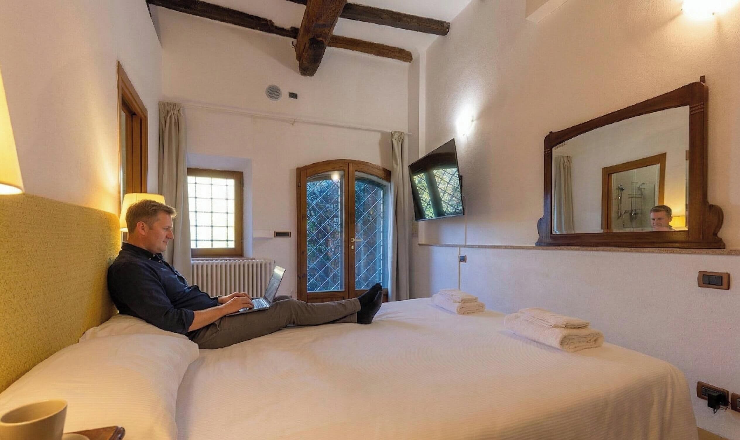 A hotel room with a bed, a TV mounted on the wall, a window with curtains, and a wooden ceiling beam.