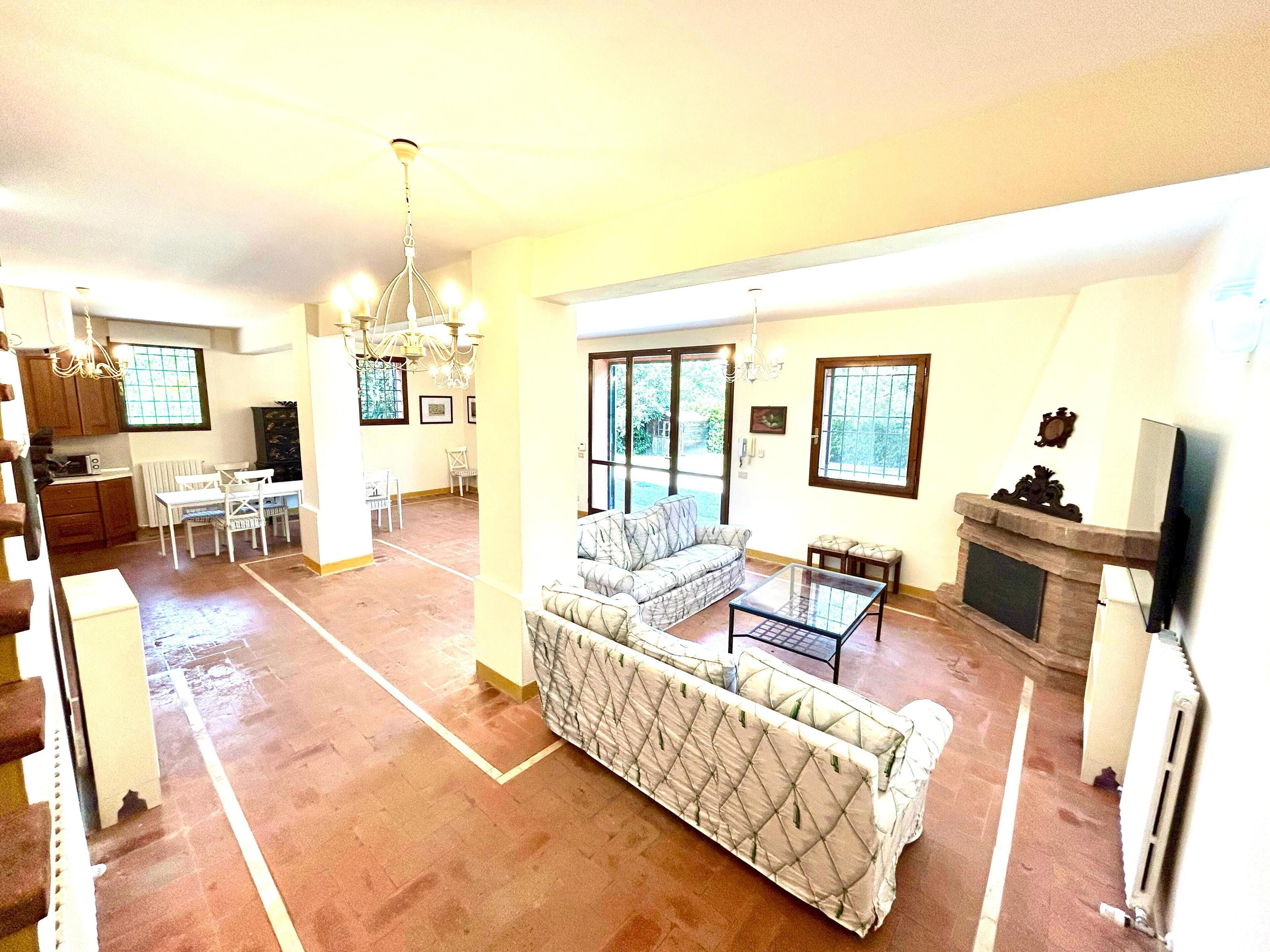 A spacious living room with a large sofa, a coffee table, and a fireplace. There is a dining area with a table and chairs, and a kitchen in the background.