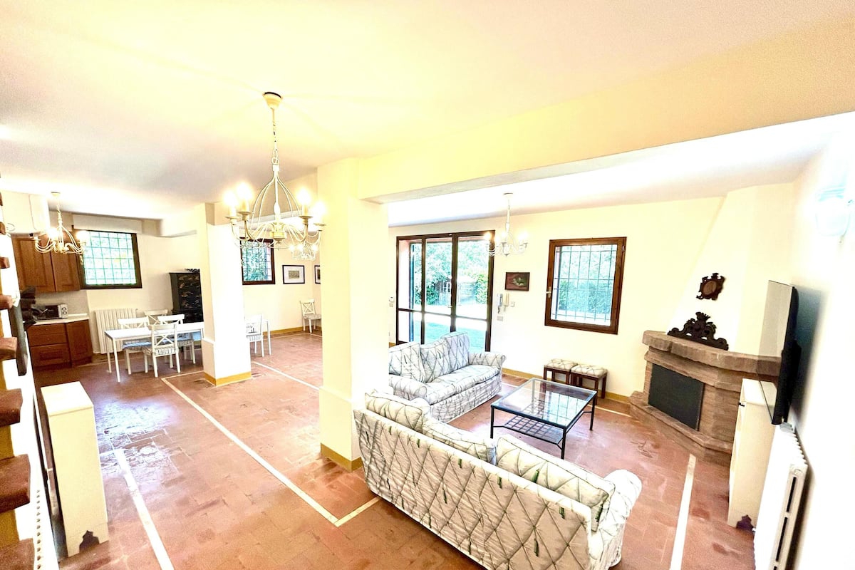 A spacious living room with a large sofa, a coffee table, and a fireplace. There is a dining area with a table and chairs, and a kitchen in the background.