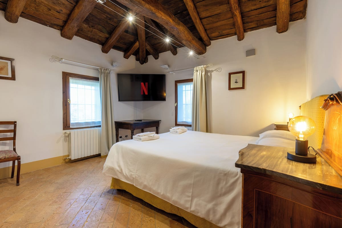 Une chambre d’hôtel avec un grand lit, des poutres apparentes en bois au plafond, un téléviseur à écran plat, une lampe de chevet et une fenêtre avec des rideaux.