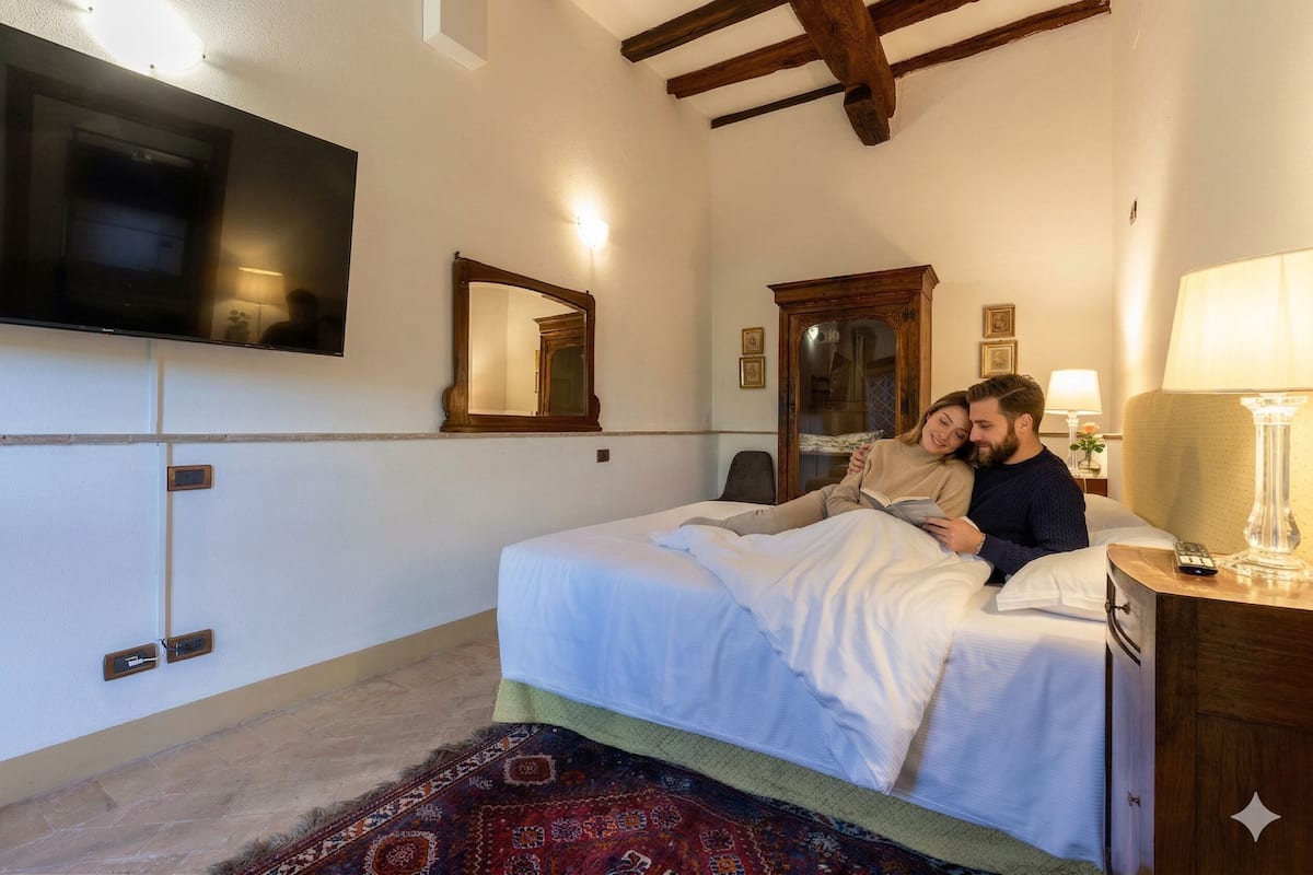 Une chambre à coucher avec un lit, un téléviseur fixé au mur, un miroir, une table de chevet avec une lampe et un tapis à motifs.