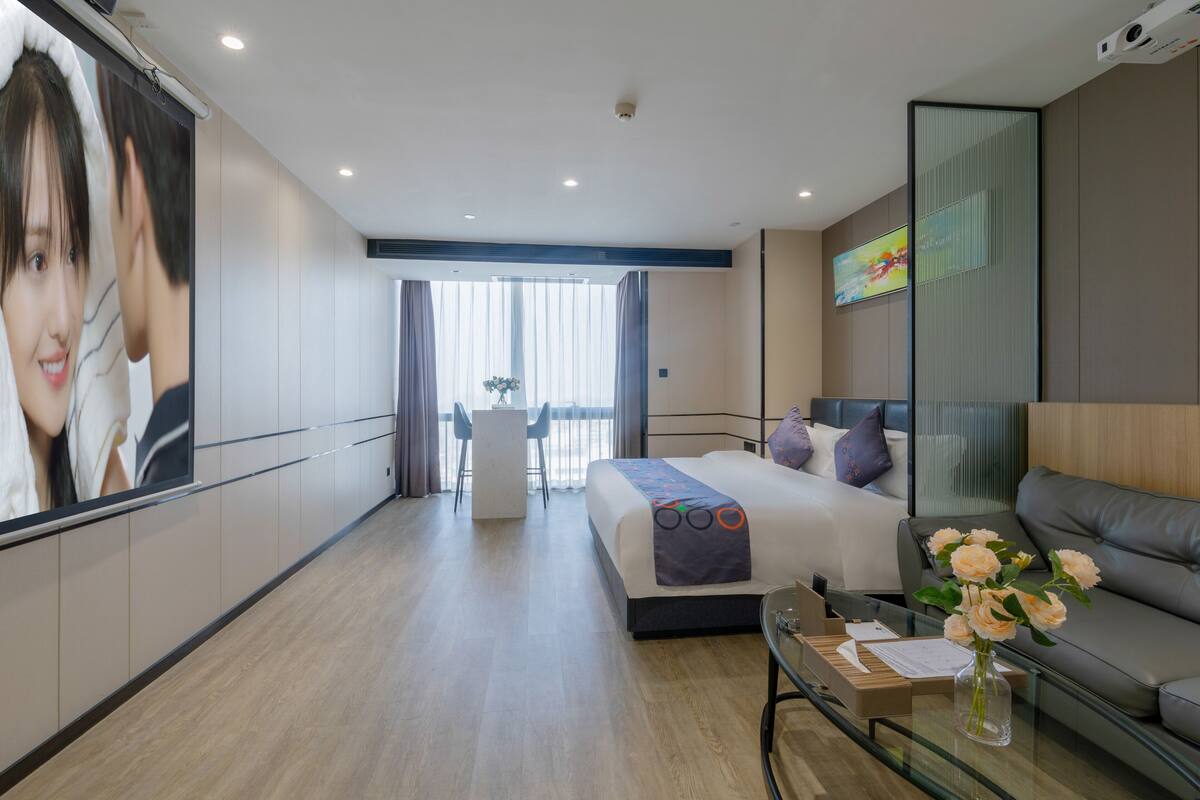 A modern hotel room with a large bed, a sofa, a glass coffee table, and a TV on the wall.