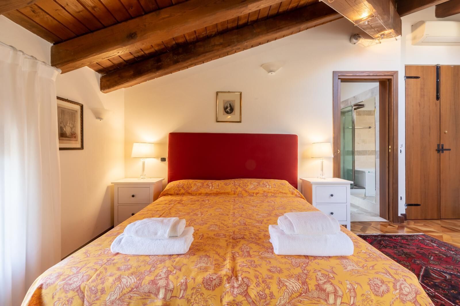 Una camera da letto con una testiera rossa, due comodini con lampade, un letto con copripiumino fantasia e una porta che dà su un bagno.