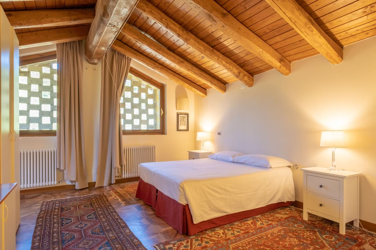 Ein Schlafzimmer mit Holzbalkendecke, einem Bett mit weißer Bettwäsche, einem Nachttisch mit Lampe und einem gemusterten Teppich.