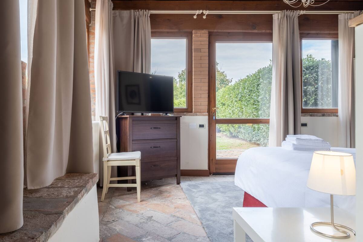 Ein Schlafzimmer mit einem Bett, einem Fernseher auf einer Holzkommode, einem kleinen Hocker und Blick ins Grüne durch das Fenster.