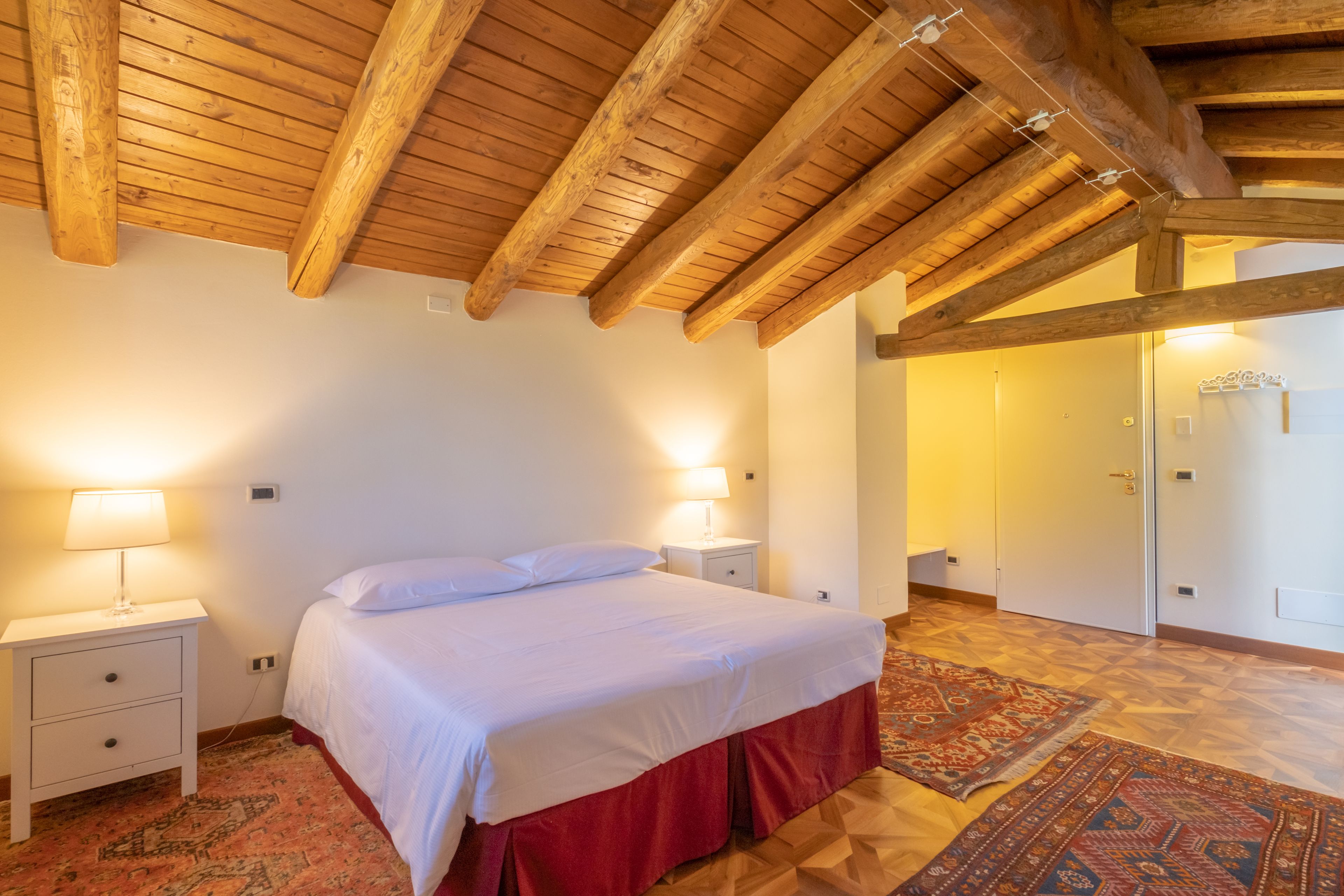 A bedroom with a wooden ceiling, a bed with white bedding, two bedside tables with lamps, and a patterned rug on the floor.
