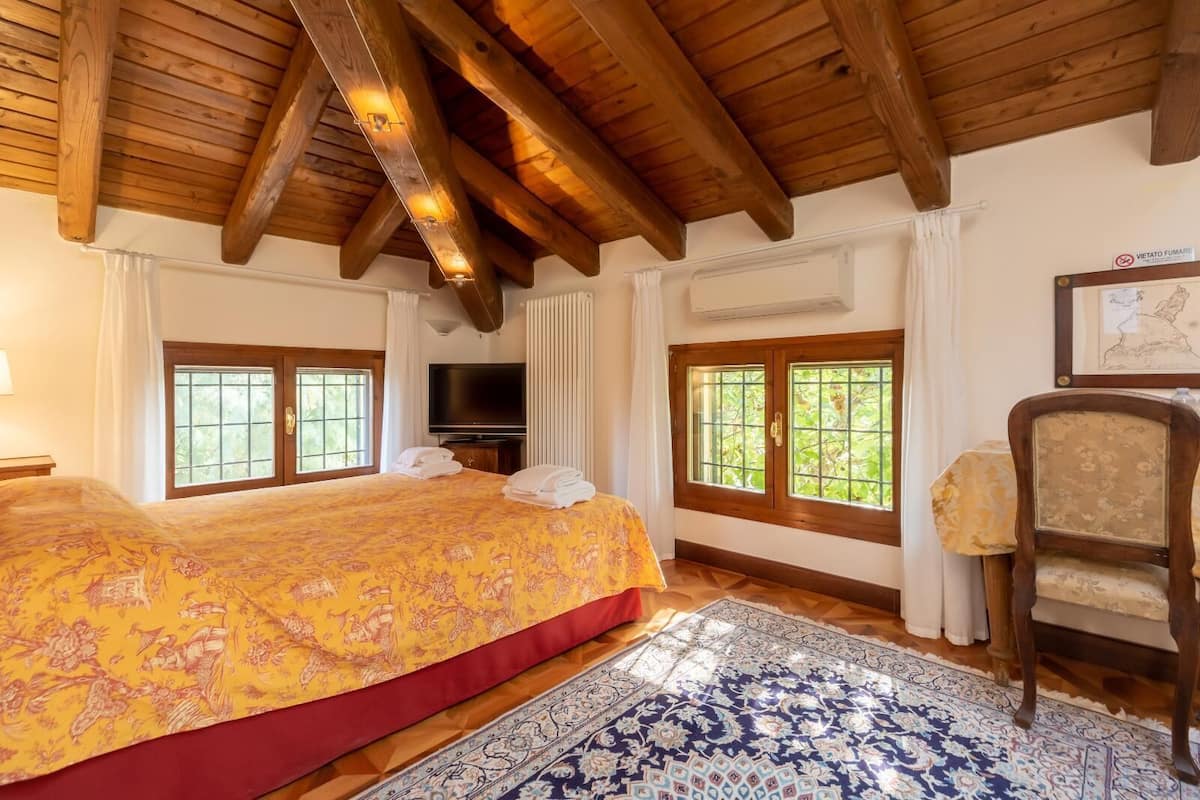 Ein Hotelzimmer mit einem großen Bett, Holzbalkendecke, einem Fernseher und einem Fenster mit Blick auf Grünflächen.