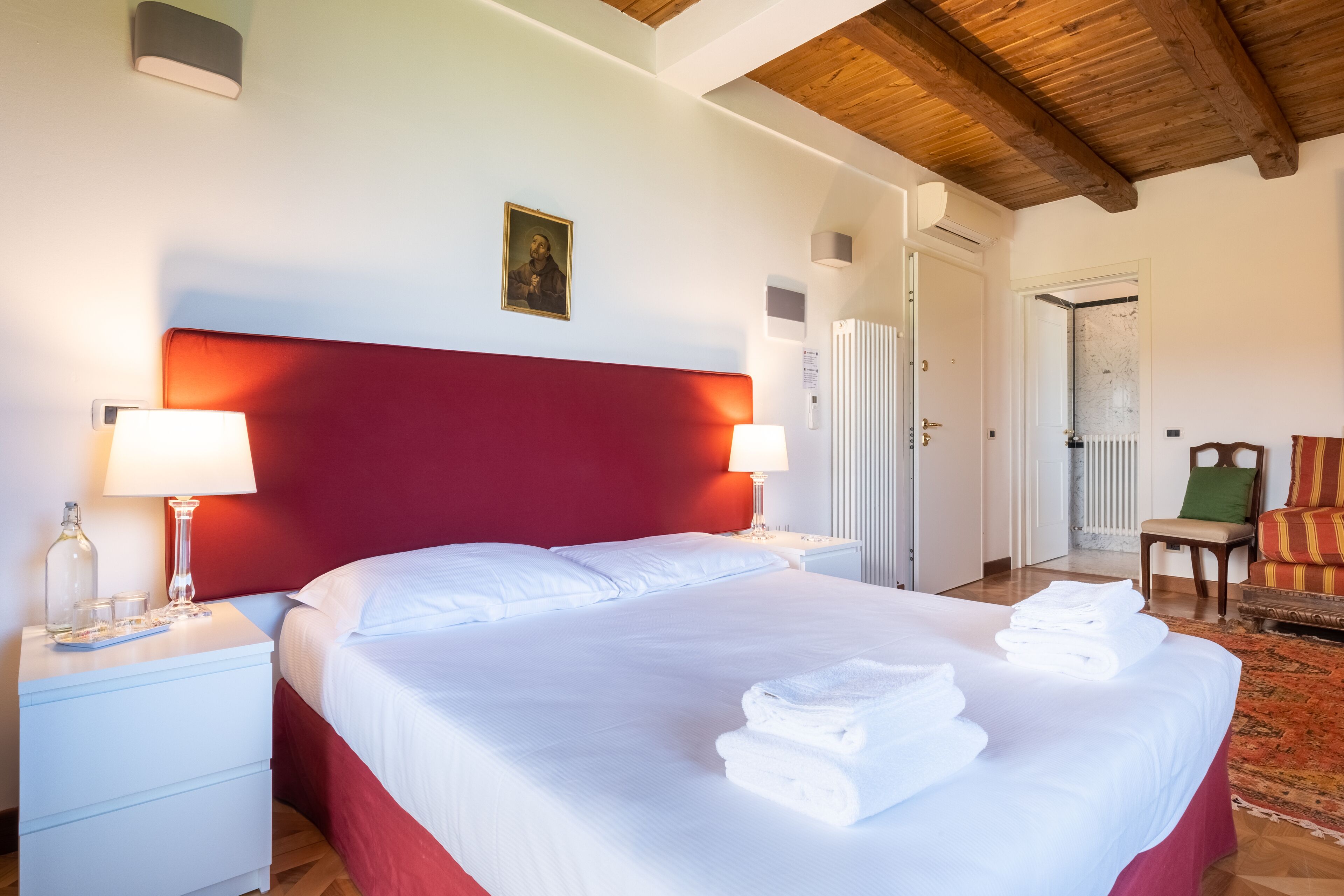 Ein Hotelzimmer mit einem großen Bett, zwei Nachttischlampen, einem roten Kopfteil, einem gerahmten Bild an der Wand, einem Sessel und einem kleinen Tisch mit einer Flasche und Gläsern.