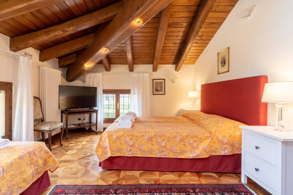 Habitación de hotel con techo de madera, cama con cabecera roja, televisor, escritorio con silla y ventana con cortinas.