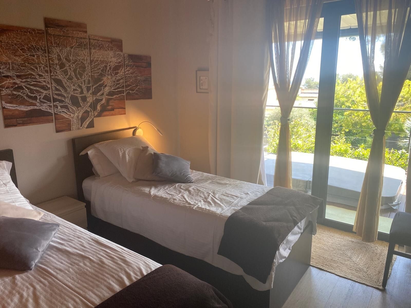 A bedroom with two beds, a nightstand, a lamp, and a view of greenery outside.