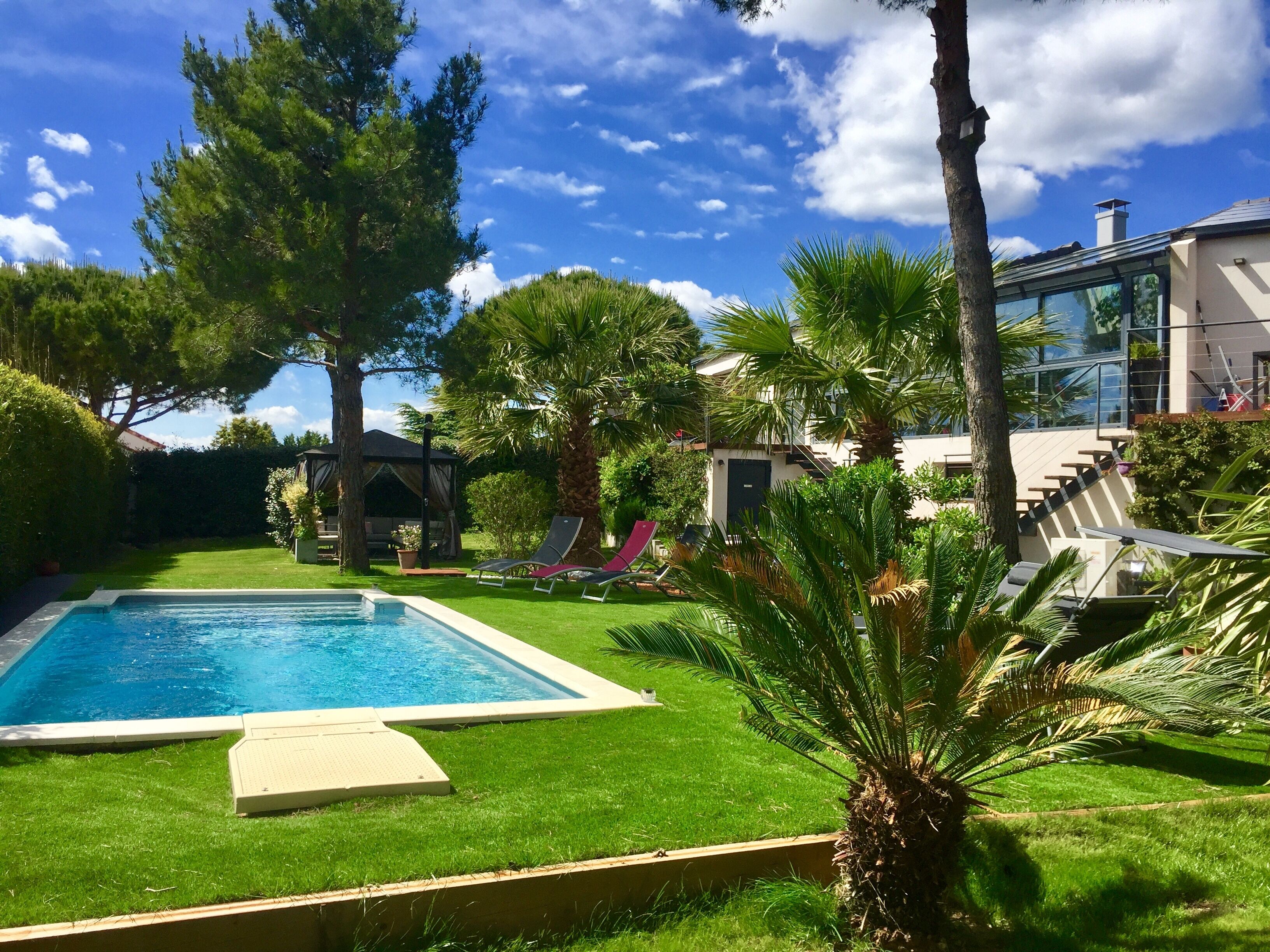 A swimming pool surrounded by lush greenery and palm trees, with a gazebo and lounge chairs in the background.