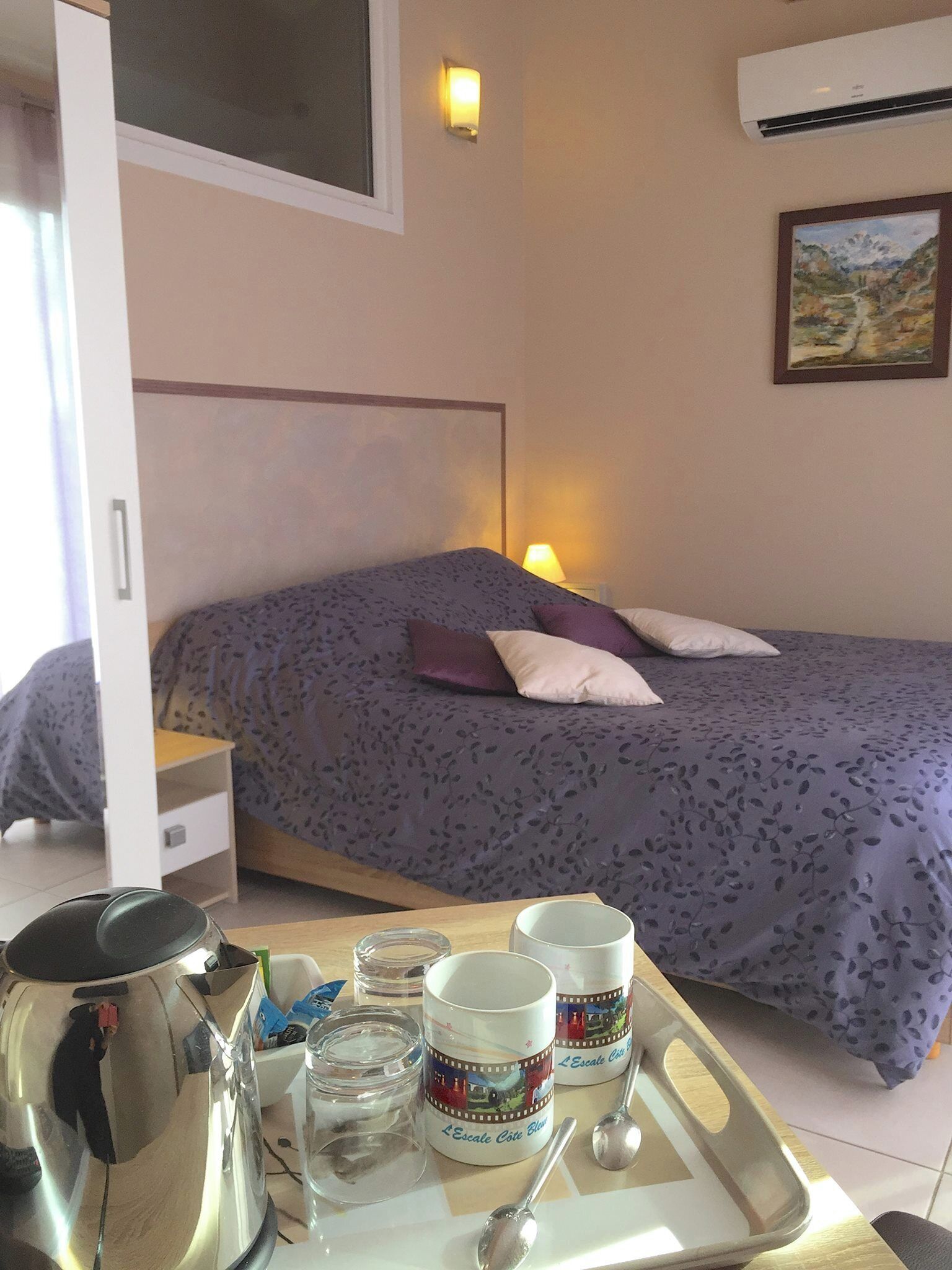 A bedroom with a bed, a bedside table, a lamp, and a tray with a kettle, cups, and jars.