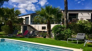 A swimming pool with lounge chairs, palm trees, and a modern building in the background. - B&B L'Escale Cote Bleue (Sausset-les-Pins)
