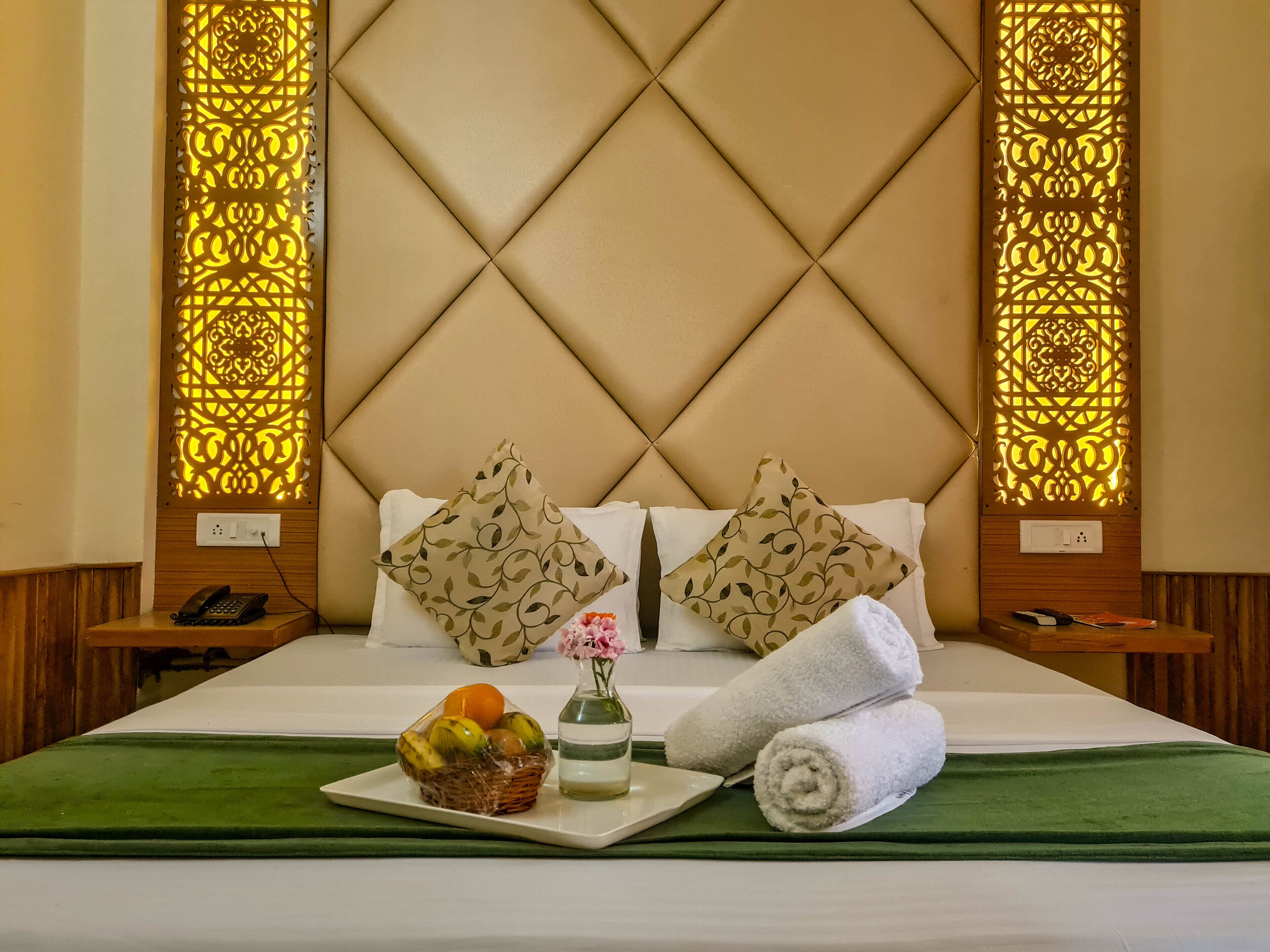 A hotel room with a tufted headboard, two decorative pillows, a bedside phone, a basket of fruit, a vase with flowers, and rolled towels on the bed.