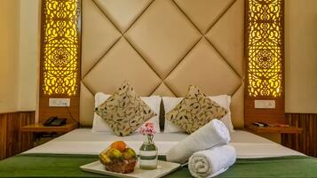 A hotel room with a tufted headboard, two decorative pillows, a bedside phone, a basket of fruit, a vase with flowers, and rolled towels on the bed.