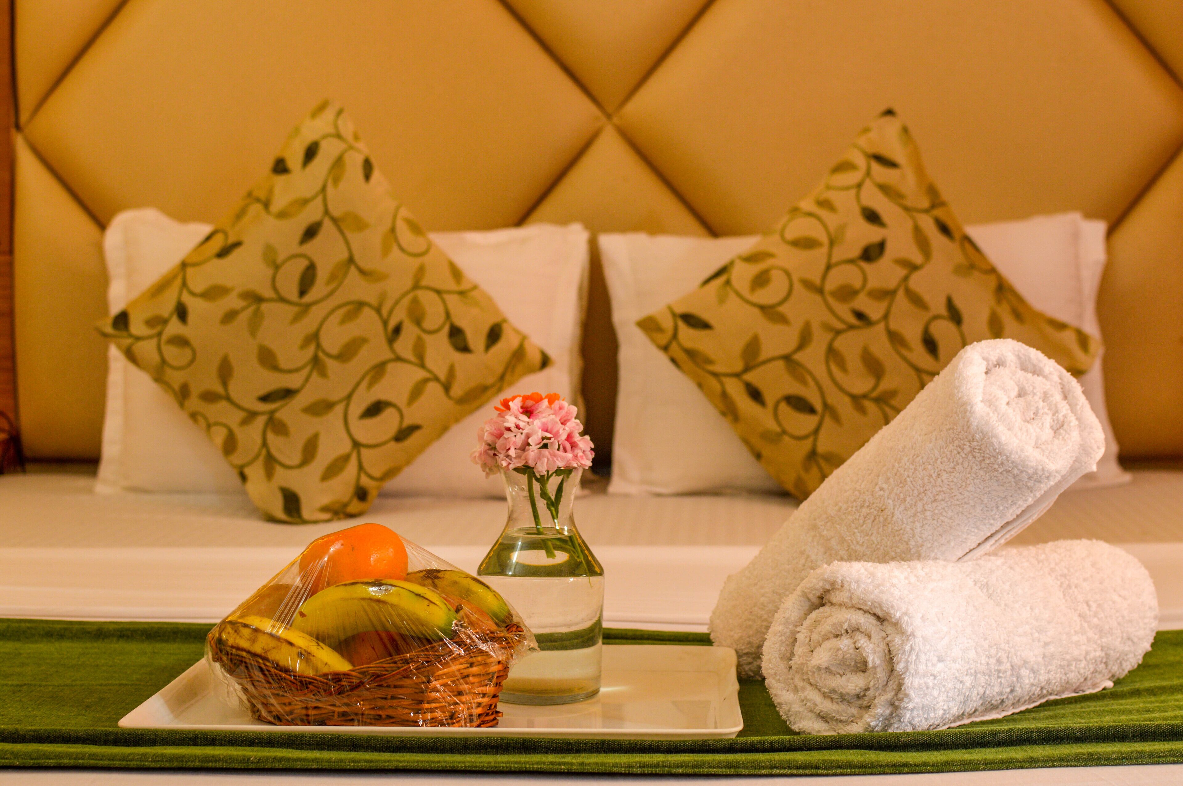 Quarto bem arrumado com cama, almofadas, cesta de frutas, vaso com flores e toalhas dobradas.