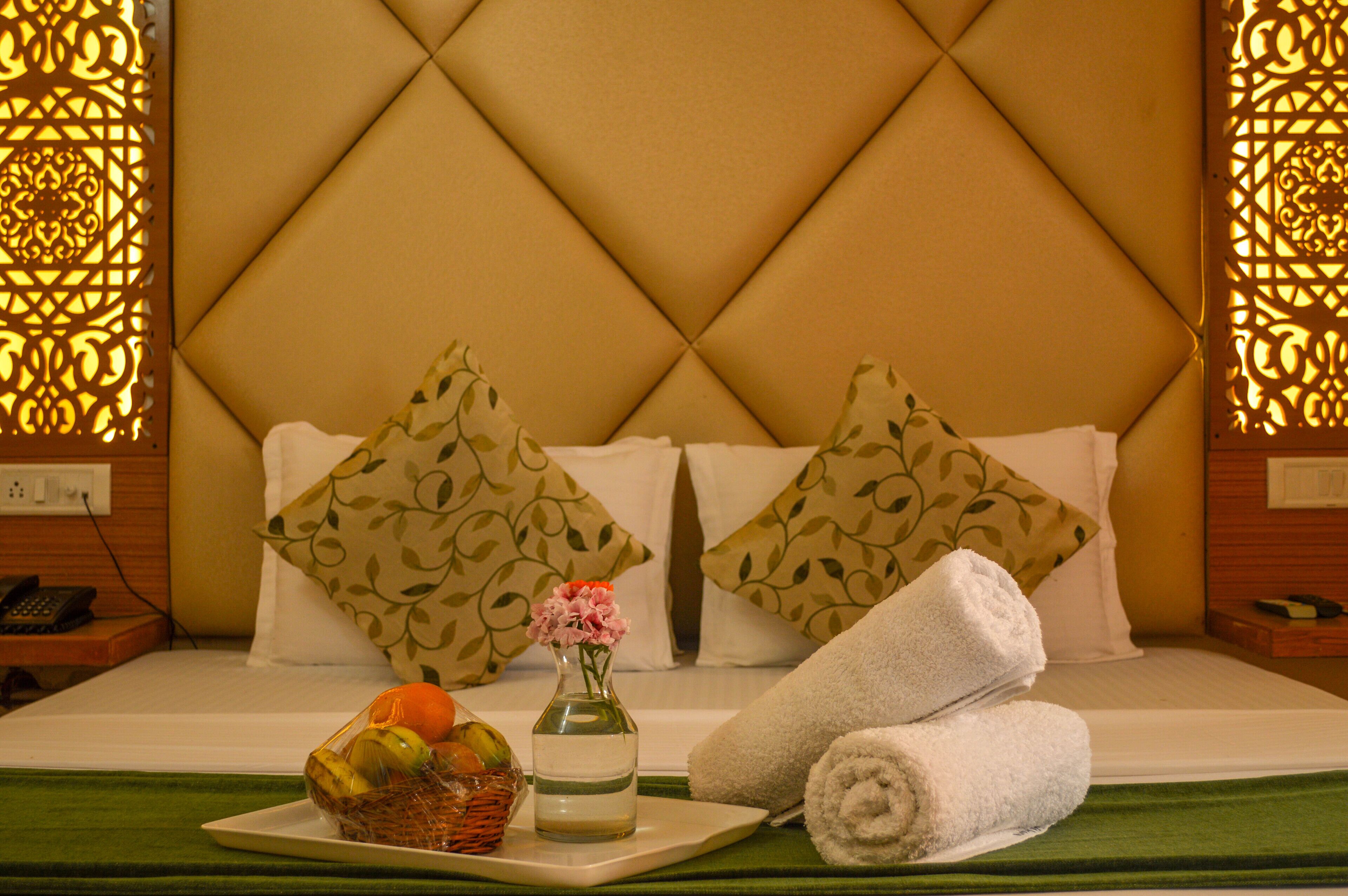 A neatly made bed with a decorative headboard, two pillows, a vase with flowers, a basket of fruit, and rolled towels.