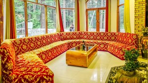 A room with a large, ornate sofa, a wooden coffee table, and large windows offering a view of a forest. - Hotel Mountain Top (Manali)