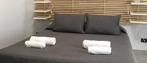 Een net opgemaakt bed met donker beddengoed en twee opgerolde handdoeken, een houten hoofdbord en nachtkastjes met lampen.