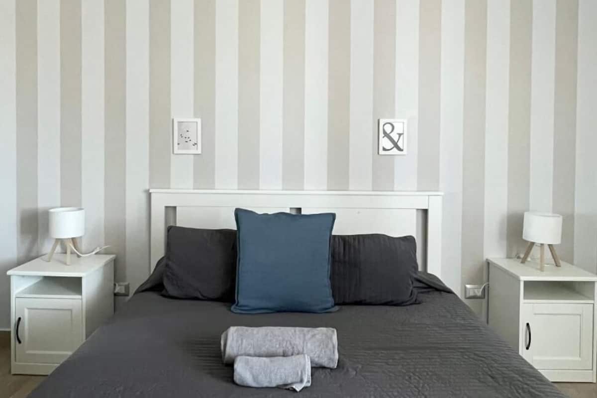 Une chambre avec un papier peint à rayures, une tête de lit blanche, deux tables de chevet avec des lampes, un lit avec une literie grise et deux oreillers.