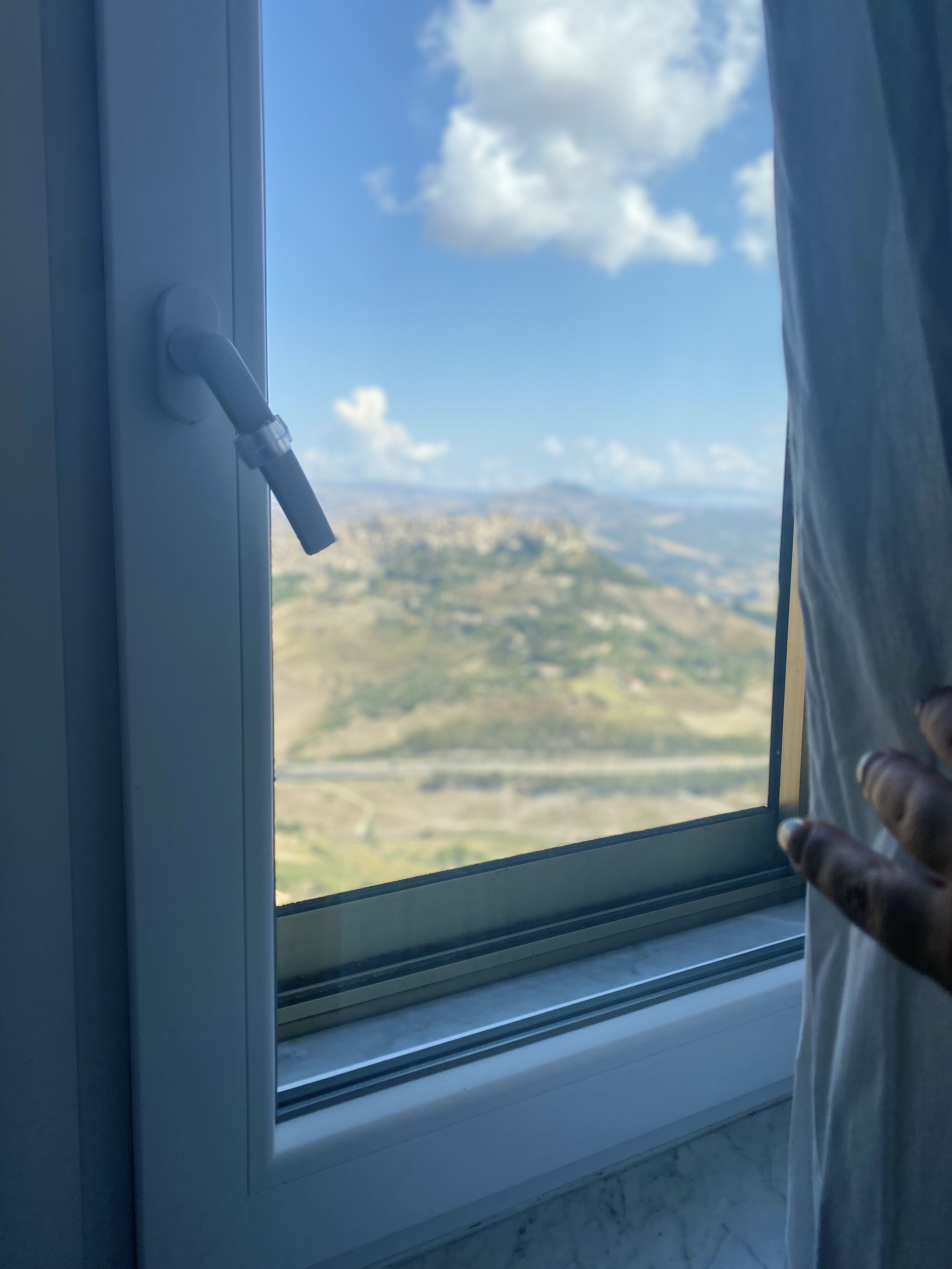 Una ventana con vista a un paisaje, una cortina entreabierta y una mano tocándola.