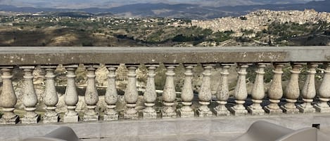 A terrace with two modern chairs, a stone balustrade, and a cityscape view under a cloudy sky.