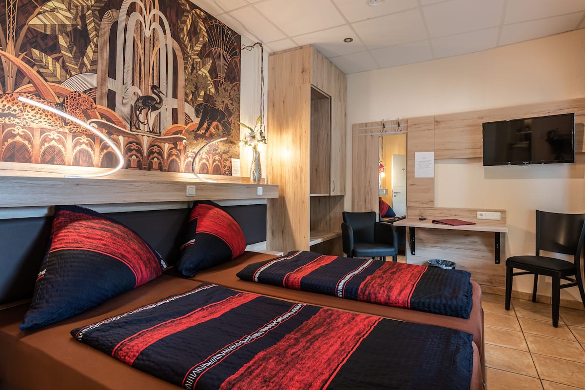 Een hotelkamer met een grote muurschildering, een bureau, twee bedden met rood en zwart beddengoed en een flatscreen televisie aan de muur.