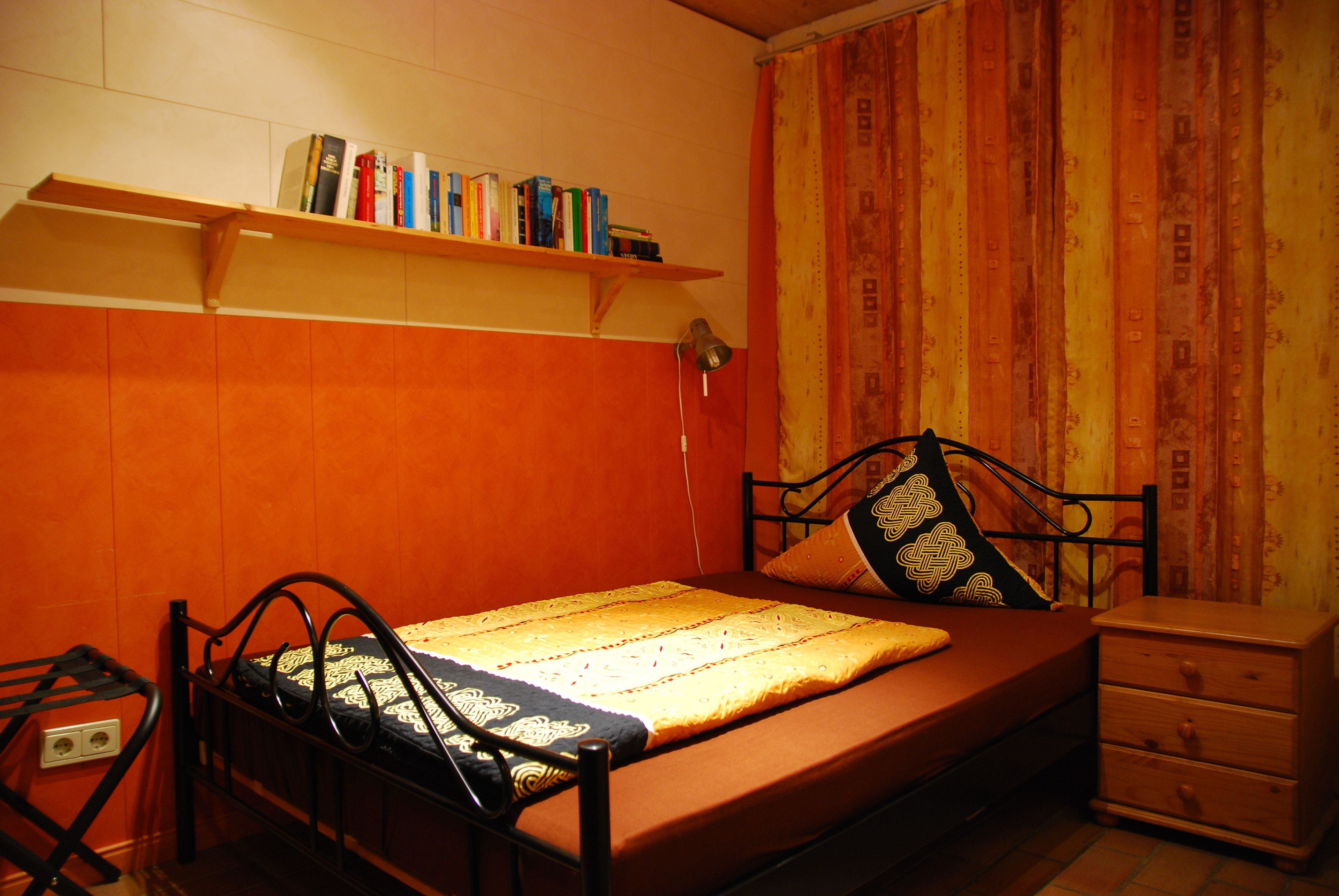 Uma cama de solteiro com estrutura de metal, mesa de cabeceira, prateleira com livros e uma cortina.