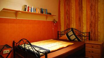 Uma cama de solteiro com estrutura de metal, mesa de cabeceira, prateleira com livros e uma cortina.