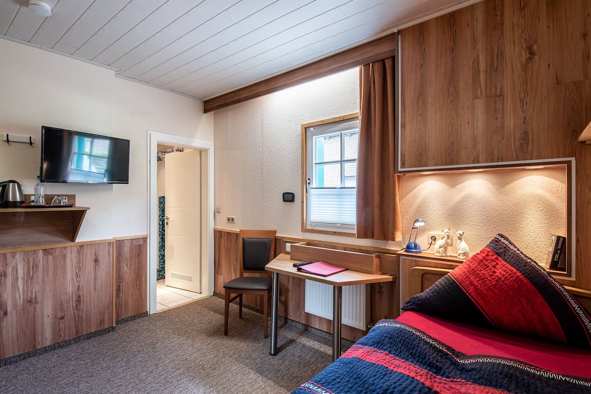 Een compacte hotelkamer met een houten bureau, een bed met een gestreept plaid, een aan de muur bevestigde televisie en een raam met gordijnen.