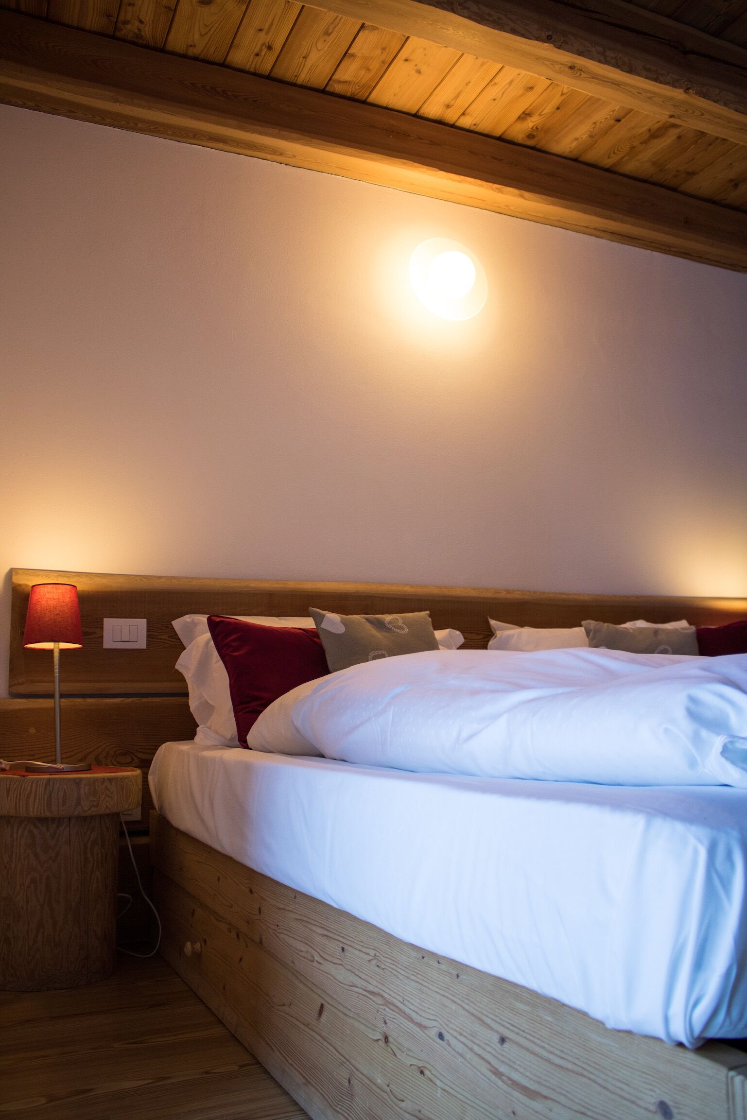 A wooden bed with white bedding, a red lamp, and a wooden nightstand.