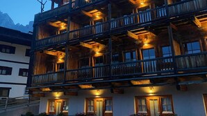 Un edificio de madera de varios pisos con balcones, una entrada bien iluminada y vistas a montañas al fondo.