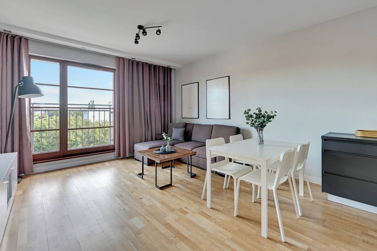 A modern living room with a sofa, dining table, and chairs, large windows with curtains, and a view of greenery outside.