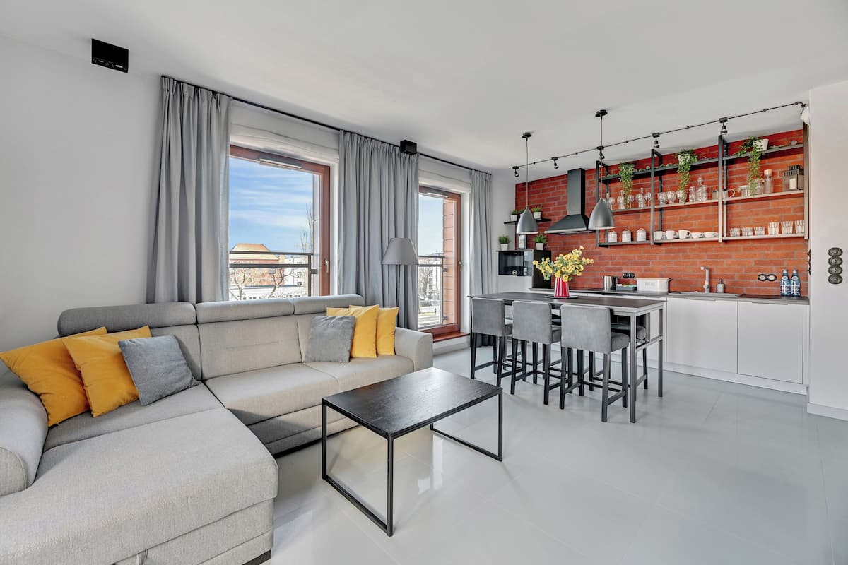 A modern living room with a grey sofa, a coffee table, and a kitchen area with a brick wall, bar stools, and a hanging light fixture.