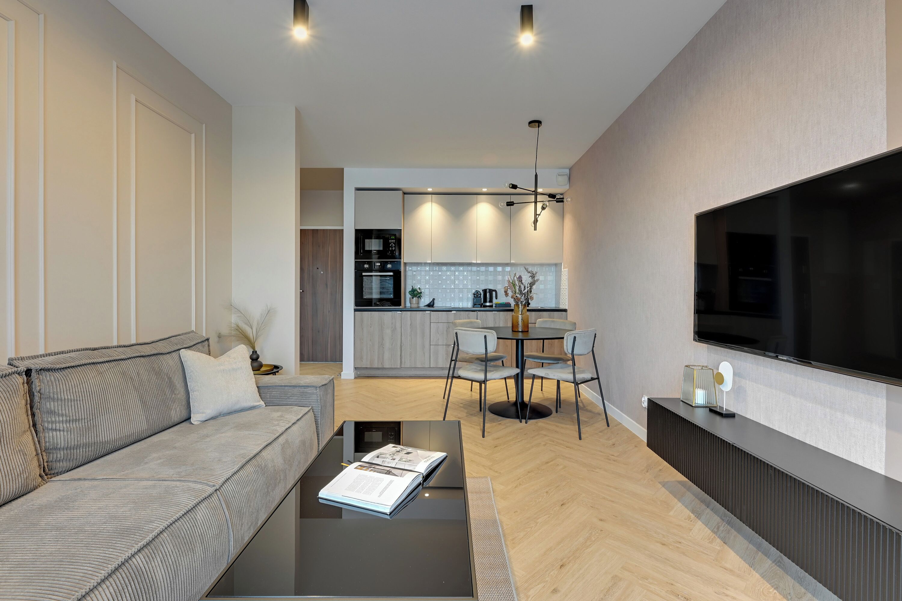 Ein modernes Wohnzimmer mit Sofa, Couchtisch und Flachbildfernseher. Ein Essbereich mit Tisch und Stühlen, und eine Küche im Hintergrund.