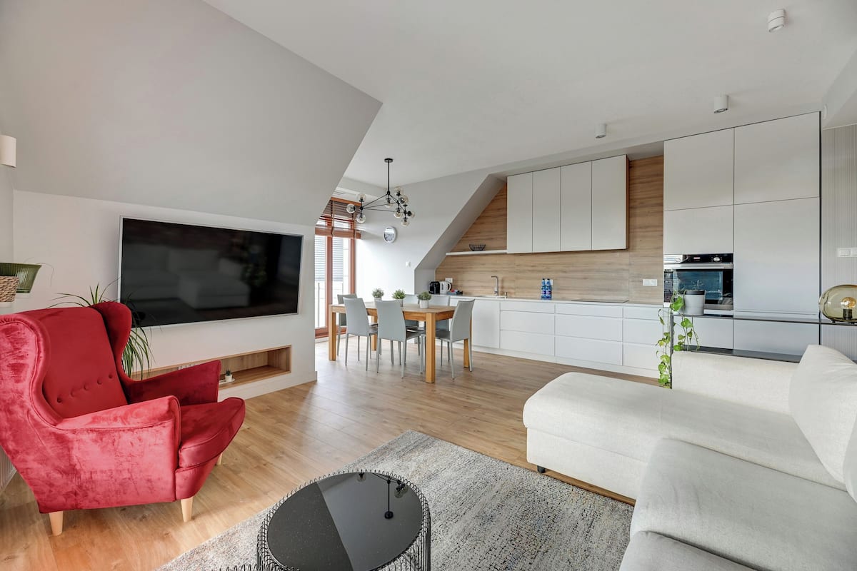 A modern living room with a red chair, white sofa, and a flat-screen TV. A dining area with a wooden table and chairs is visible in the background.