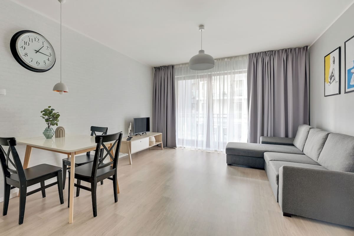 A modern living room with a dining area, a grey sofa, a TV on a stand, and a large window with curtains.