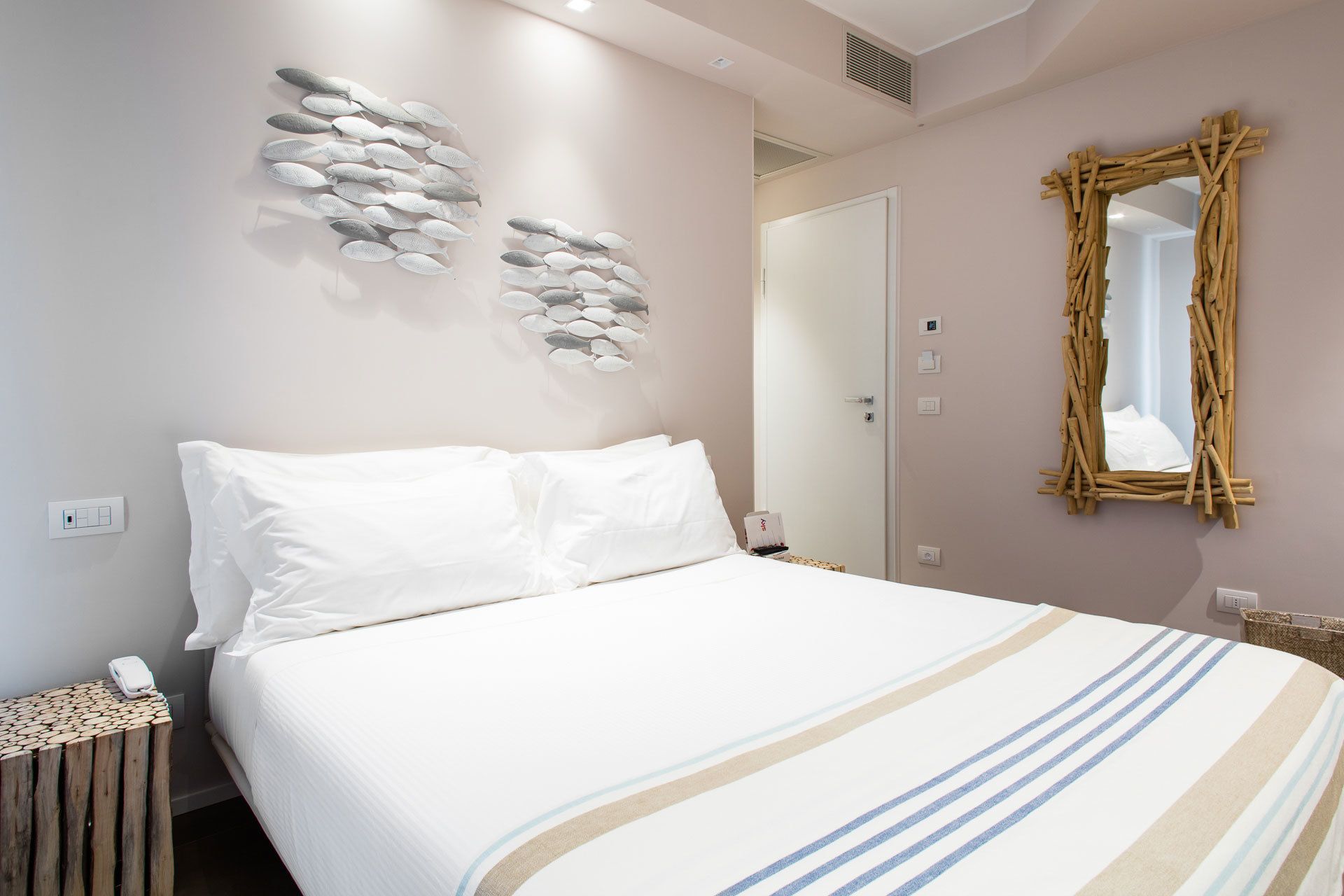 Una cama bien hecha con ropa de cama blanca y una manta a rayas, un espejo montado en la pared y una obra de arte con piedras apiladas.