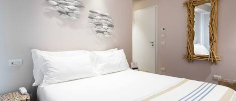 Una cama bien hecha con ropa de cama blanca y una manta a rayas, un espejo montado en la pared y una obra de arte con piedras apiladas.