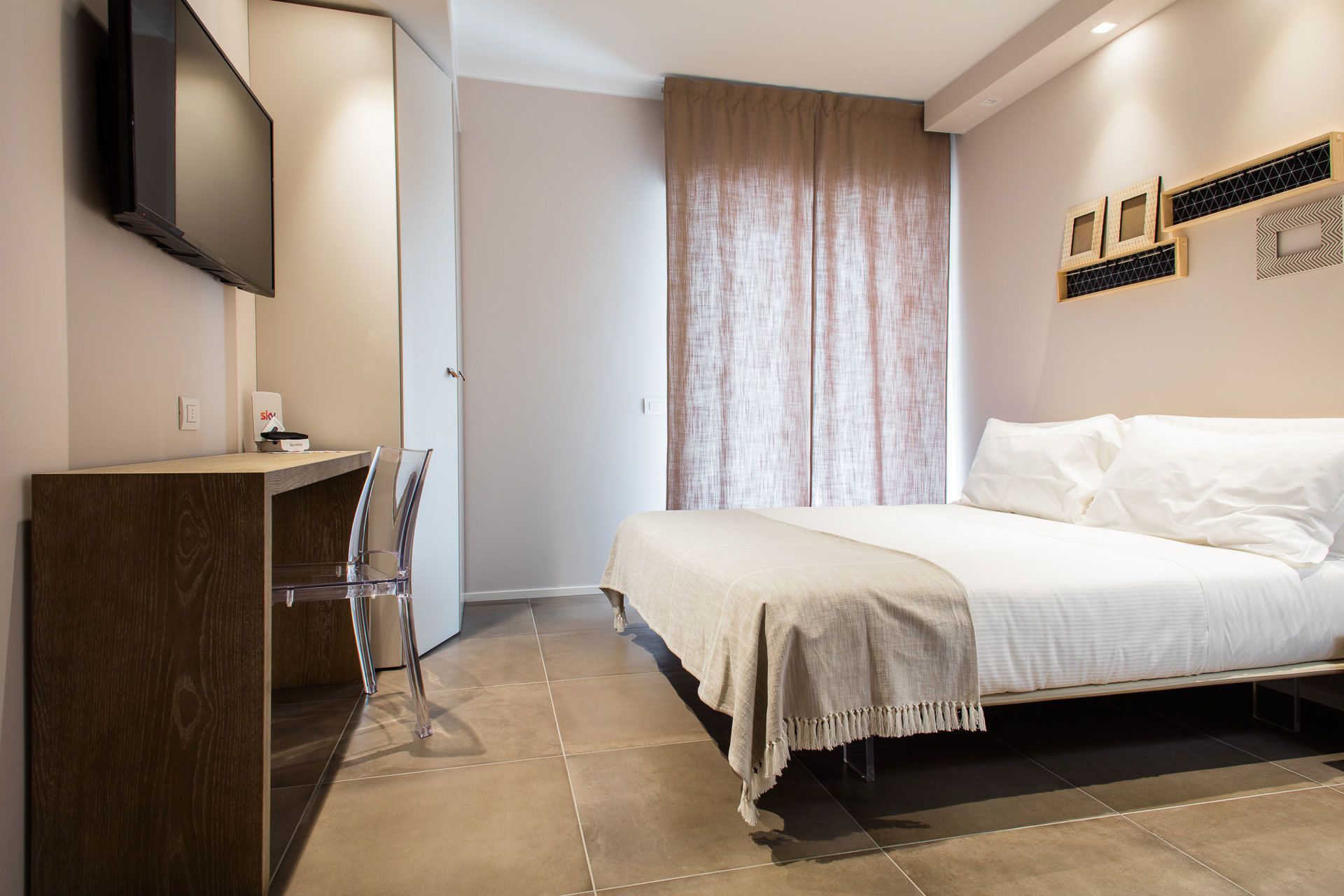 A bedroom with a bed, a desk, a chair, a TV, and a window with curtains.