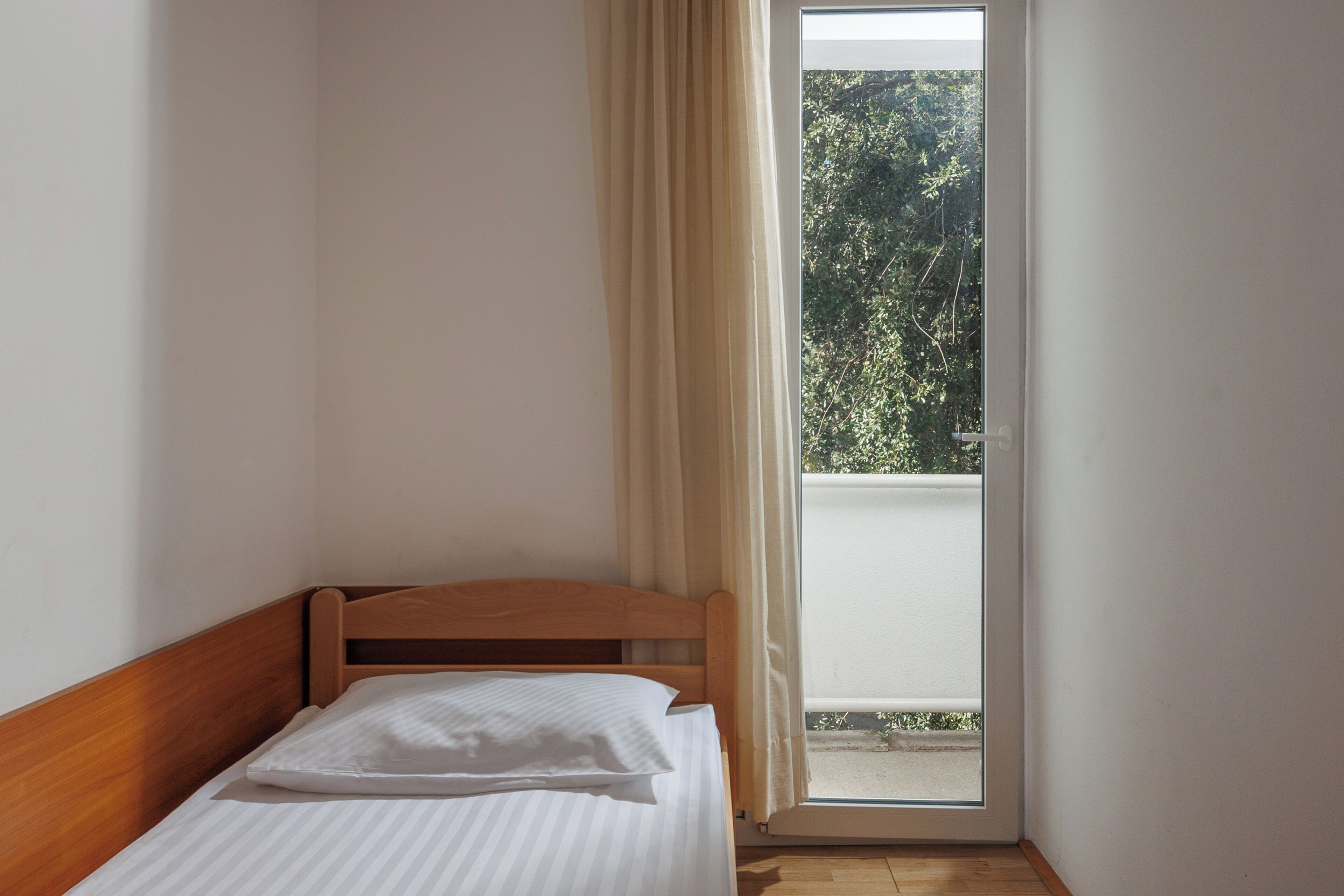 Una habitación pequeña con cama individual, cabecera de madera, ventana con cortinas y vistas a árboles.