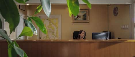 A reception desk with a wooden counter, a framed landscape painting, a map on the wall, and a potted plant in the foreground.