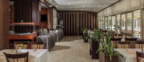 A restaurant with a long counter, multiple tables, and chairs. There are potted plants and a large window with a view.