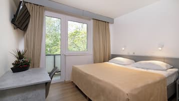 Een slaapkamer met een bed, een bureau, een potplant, een raam met gordijnen en uitzicht op bomen.